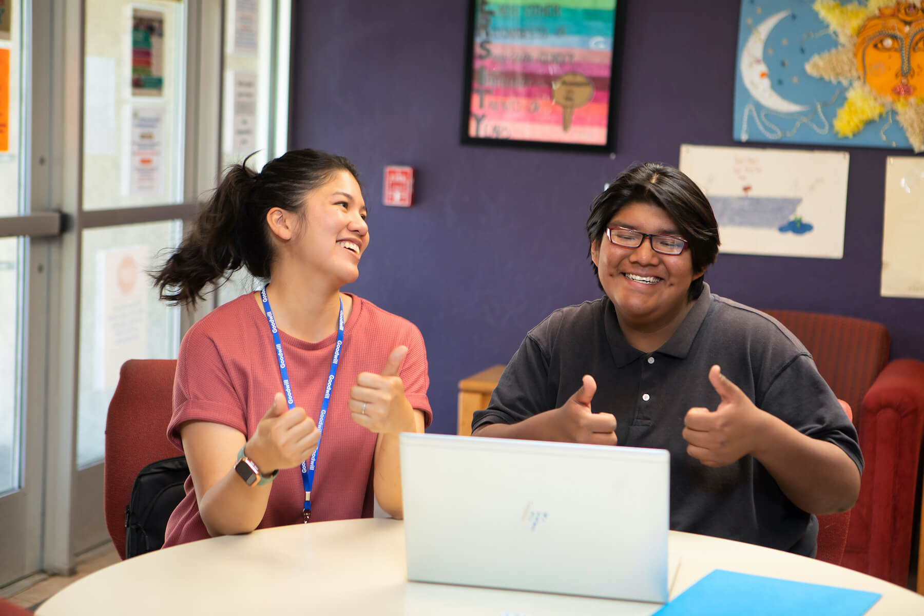 A teacher and student work together at a table with a laptop computer. They are giving a thumbs up.