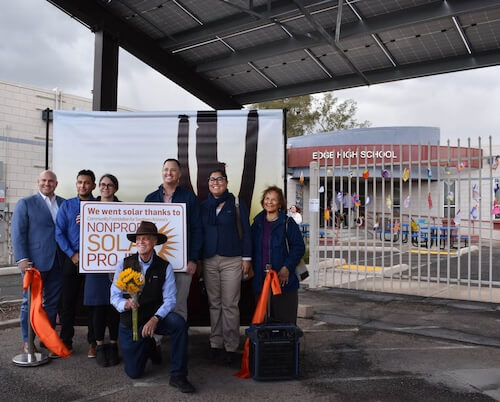 A group of people stand in front a sign announcing a solar project. Edge High School is in the background.