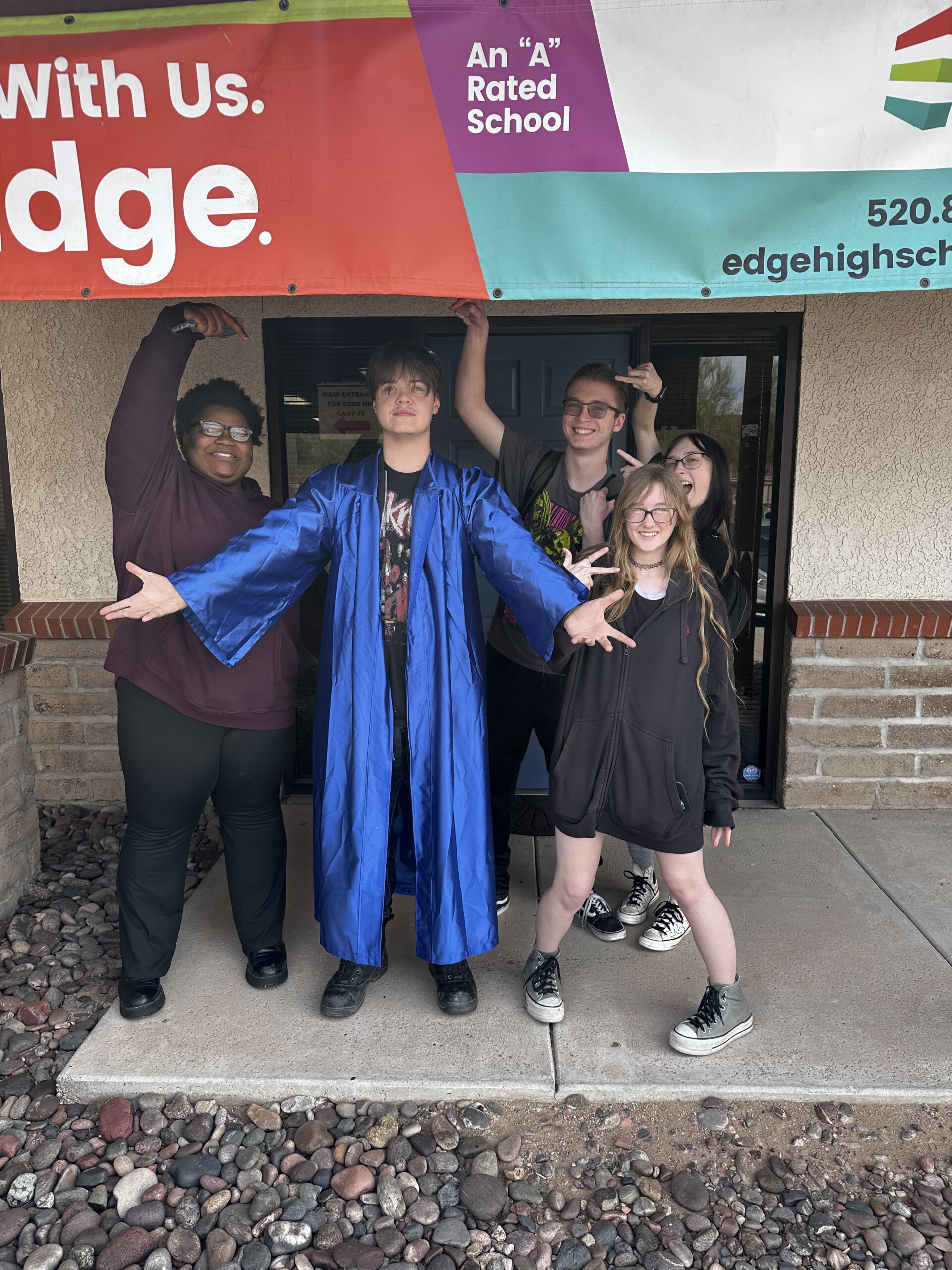Several Edge High School students stand in front of the main doorway. One is in a blue graduation gown with arms stretched out in front of them.